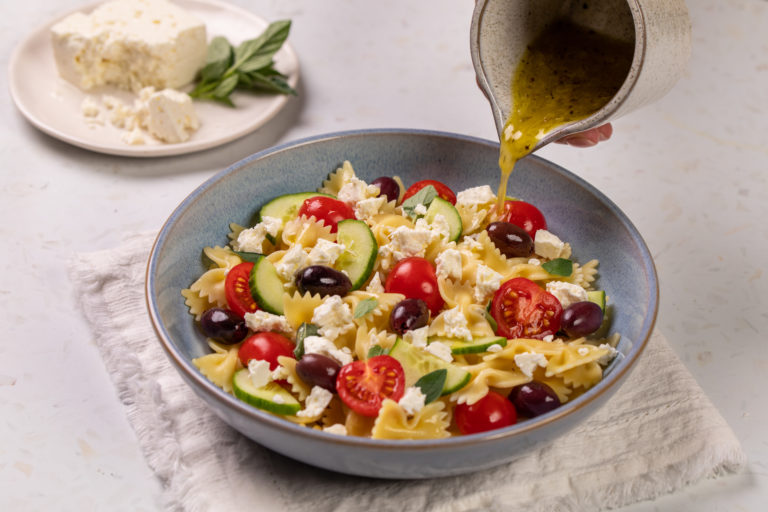 Bowl of greek salad with feta, olives, tomatoes, cucumbers, feta cheese, pasta. Drizzle of salad dressing on top.