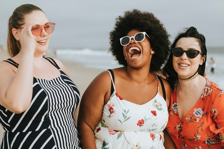 Two white and one black plus size women at the beach, smiling and laughing. 