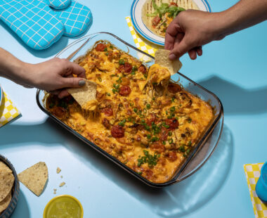 Hands dipping chips into a tray of cheese corn chicken dish