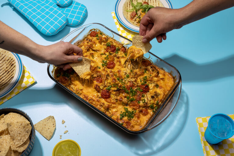 Hands dipping chips into a tray of cheese corn chicken dish